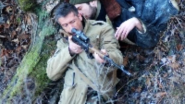 Zwei Männer liegen im Unterholz, einer zielt mit einem Gewehr.