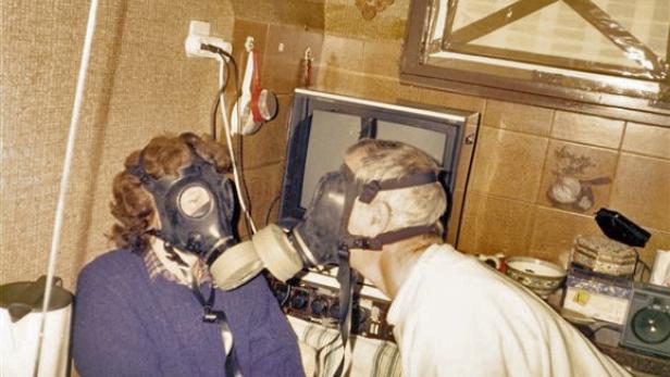 Zwei Personen mit Gasmasken stehen vor einem alten Fernseher.