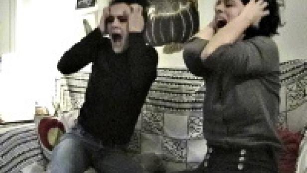 Zwei Personen sitzen auf einem Sofa und schreien mit den Händen an den Ohren.