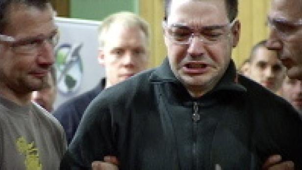 Ein Mann mit Schutzbrille verzieht schmerzhaft das Gesicht, umringt von anderen Männern.