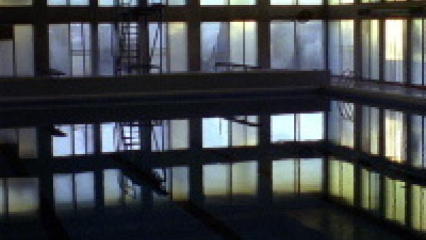 Ein leeres Schwimmbecken mit Sprungturm spiegelt sich im Wasser.