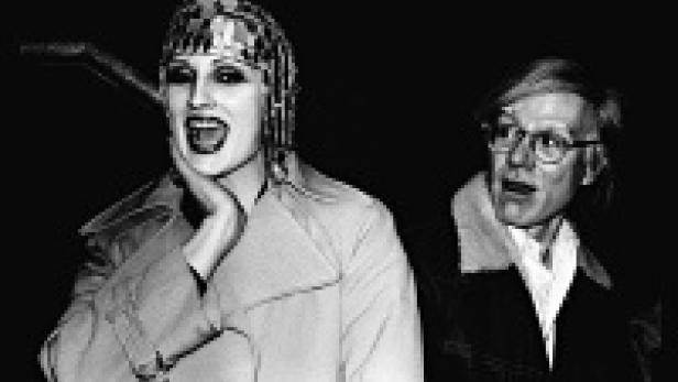 Eine Frau mit Kopfschmuck und Trenchcoat steht neben Andy Warhol.