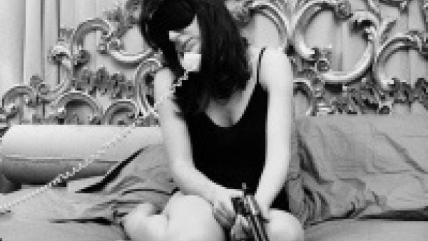 Eine Frau sitzt mit verbundenen Augen auf einem Bett, telefoniert und hält eine Pistole in der Hand.