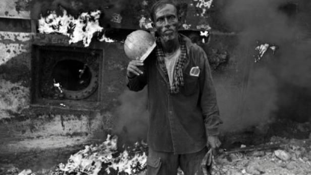 Ein erschöpfter Arbeiter hält einen Helm vor einem zerstörten, rauchenden Gebäude.