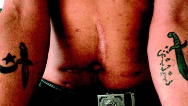 Ein Mann mit Tätowierungen und einer Narbe auf der Brust trägt einen Gürtel.