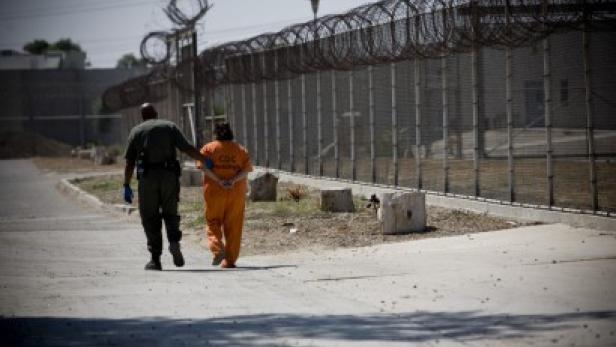 Ein Gefängniswärter begleitet einen Gefangenen in oranger Kleidung entlang eines Zauns mit Stacheldraht.