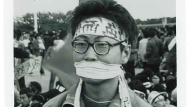 Ein junger Mann mit Brille und Stirnband hat einen Knebel im Mund, inmitten einer Menschenmenge.