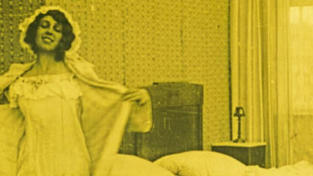 Eine Frau im Nachthemd steht lächelnd neben einem Bett.