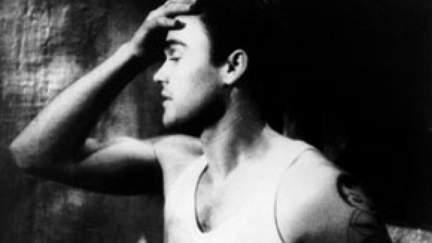 Ein Mann mit Tätowierung und Unterhemd hält sich die Hand an die Stirn.