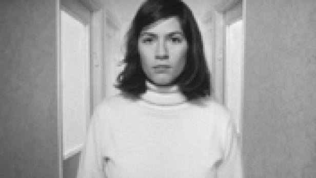 Eine Frau mit dunklen Haaren und einem weißen Rollkragenpullover steht in einem Flur.