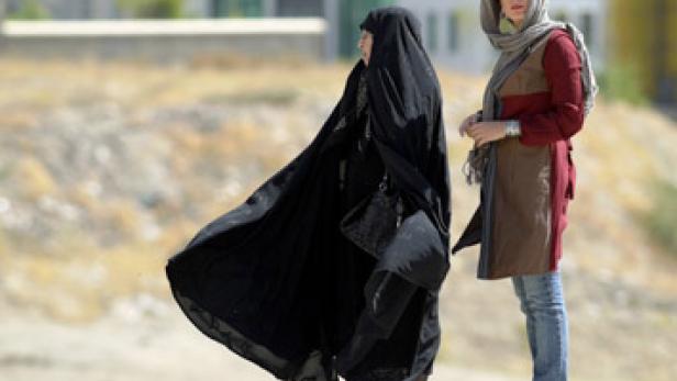 Zwei Frauen stehen im Freien, eine in traditioneller schwarzer Kleidung, die andere in moderner Kleidung.
