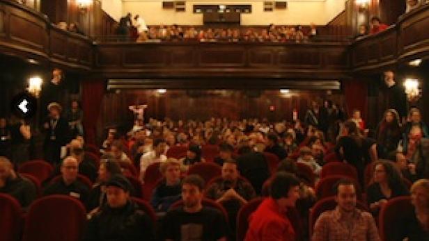 Ein volles Theater mit Zuschauern auf den Sitzen und auf dem Balkon.