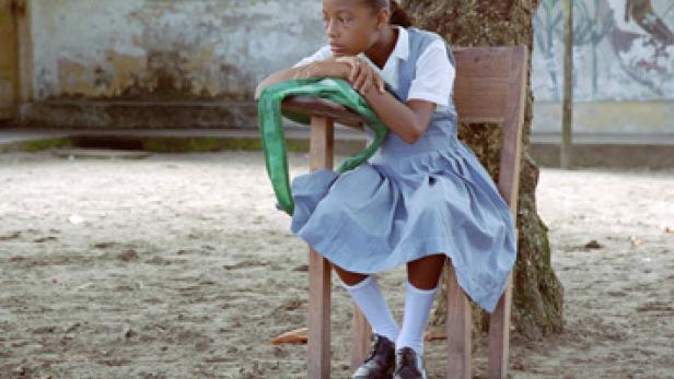 Ein junges Mädchen in Schuluniform sitzt auf einem Stuhl und blickt nachdenklich zur Seite.