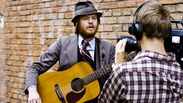 Ein Musiker mit Hut und Gitarre wird von einem Kameramann gefilmt.