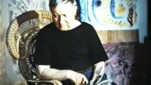 Ein älterer Mann sitzt in einem Korbstuhl vor einem abstrakten Gemälde.