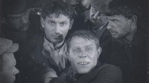 Eine Gruppe von Männern, einige mit besorgten Gesichtsausdrücken, in einer dunklen Umgebung.
