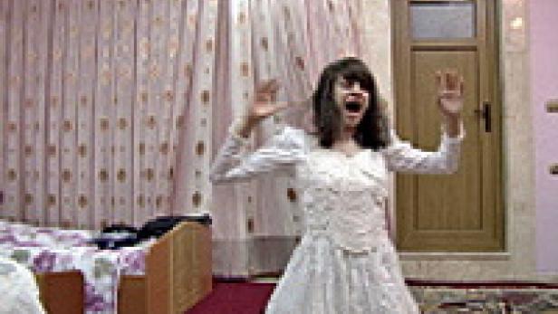 Eine Person im weißen Kleid steht mit erhobenen Händen und geöffnetem Mund in einem Raum.