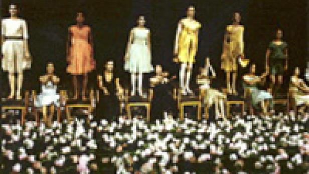 Eine Modenschau mit Models in farbenfrohen Kleidern, die auf Stühlen vor einem großen Publikum sitzen.