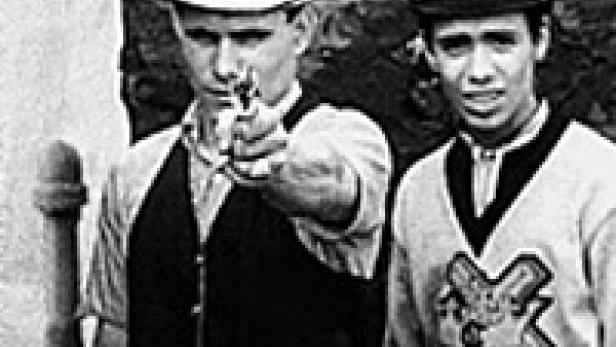 Zwei junge Männer, einer mit einer Pistole, posieren für ein Schwarzweißfoto.