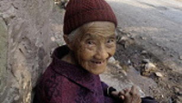 Eine ältere Frau mit roter Mütze sitzt an einer Wand und lächelt.