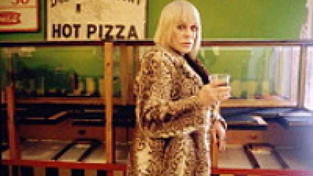 Eine Frau mit blonden Haaren und einem gemusterten Mantel hält ein Glas in der Hand.