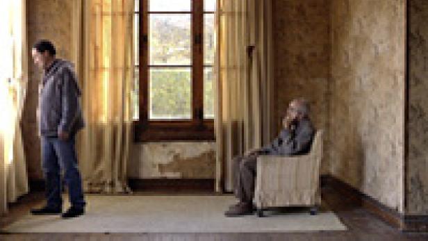 Ein Mann steht am Fenster, während ein älterer Mann in einem Sessel sitzt.