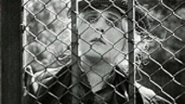 Eine Frau mit Mütze blickt durch einen Maschendrahtzaun nach oben.