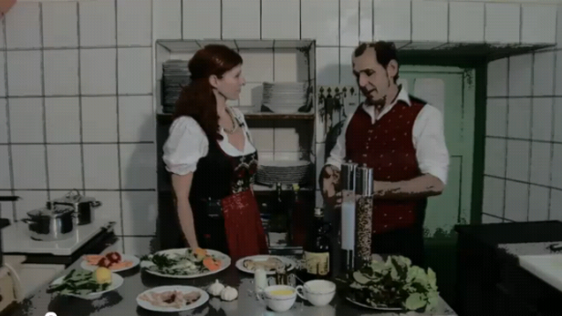 Ein Mann und eine Frau in Tracht stehen in einer Küche und bereiten Speisen zu.