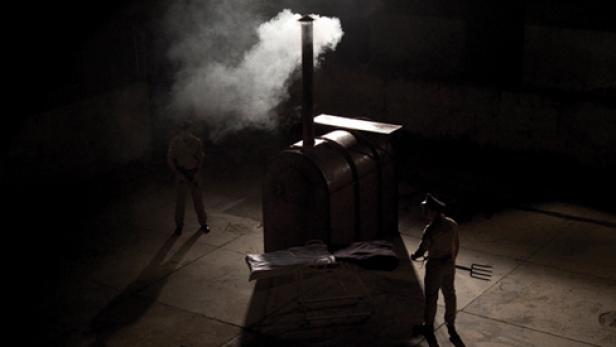Zwei uniformierte Männer stehen im Dunkeln neben einem Ofen, aus dem Rauch aufsteigt.