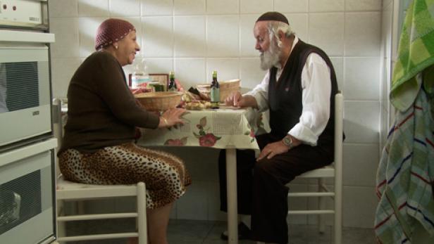 Ein älteres jüdisches Paar sitzt an einem Tisch in einer Küche und unterhält sich.