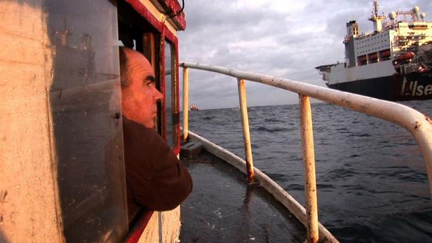 Ein Mann blickt von einem kleinen Boot auf ein großes Schiff auf dem Meer.