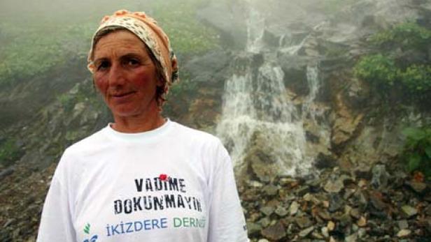 Eine Frau mit Kopftuch und einem T-Shirt mit der Aufschrift „VADIME DOKUNMAYIN“ vor einem Wasserfall.