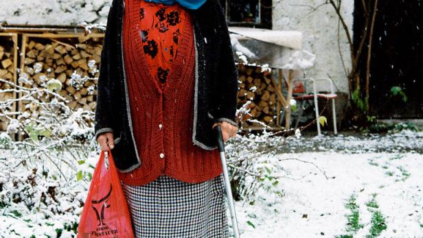 Eine ältere Frau mit Kopftuch und Stock steht im Schnee vor einem Haus.