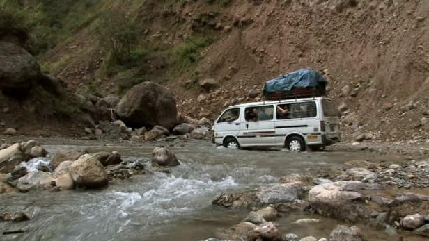 Ein weißer Van fährt durch einen Fluss in einer bergigen Landschaft.