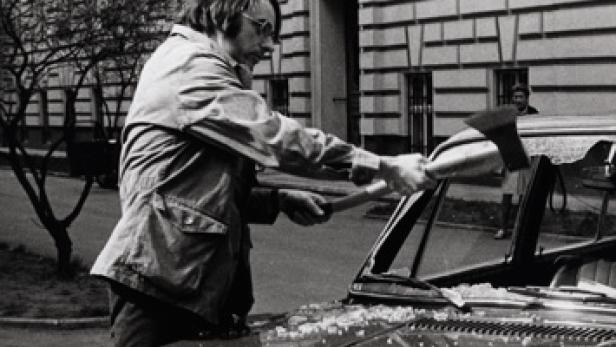 Ein Mann mit Brille zertrümmert mit einer Axt die Windschutzscheibe eines Autos.