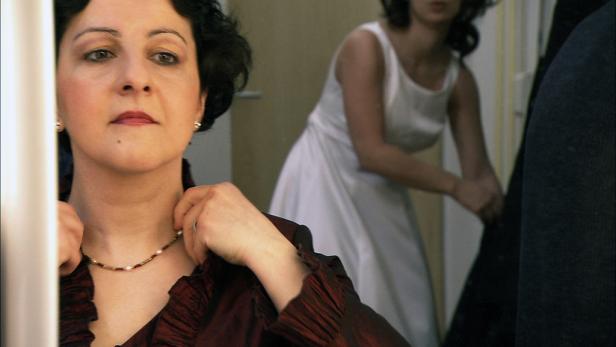 Eine Frau legt eine Halskette an, während eine andere Frau im Hintergrund ein weißes Kleid trägt.