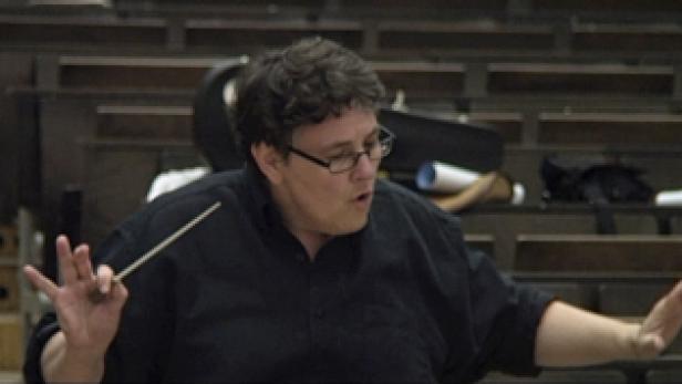 Ein Dirigent mit Brille dirigiert ein Orchester.