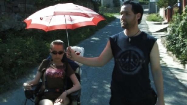 Ein Mann schützt eine Frau im Rollstuhl mit einem rot-weißen Sonnenschirm vor der Sonne.