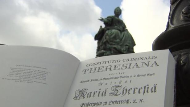 Ein aufgeschlagenes Buch mit dem Titel „Constitutio Criminalis Theresiana“ vor einem Denkmal von Maria Theresia.