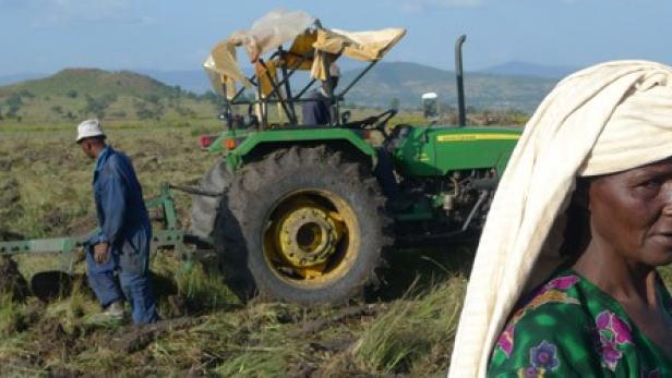 Eine Frau mit Kopftuch steht auf einem Feld, während ein Mann mit einem Traktor pflügt.