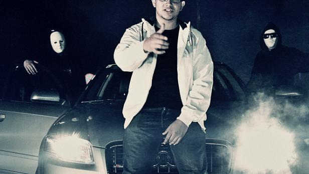 Ein junger Mann posiert vor einem Auto, flankiert von zwei Personen mit Masken.