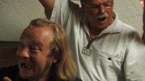 Zwei Männer, einer mit langen blonden Haaren und einer mit Brille, scheinen ausgelassen zu feiern.