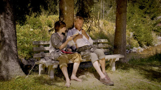 Ein Junge und ein älterer Mann sitzen barfuß auf einer Bank im Grünen.