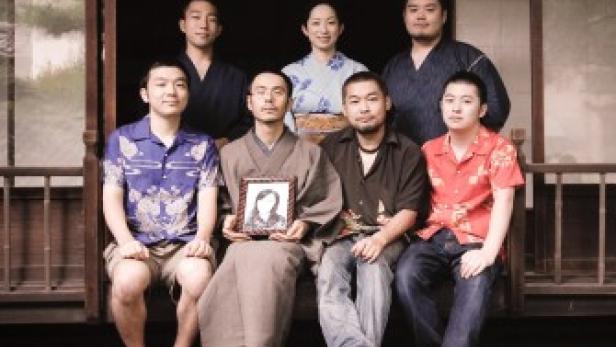 Eine Gruppe von sieben Personen posiert vor einem traditionellen japanischen Haus, eine Person hält ein Porträt.