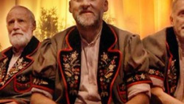 Drei ältere Männer in Trachten sitzen nebeneinander vor einem Vorhang.