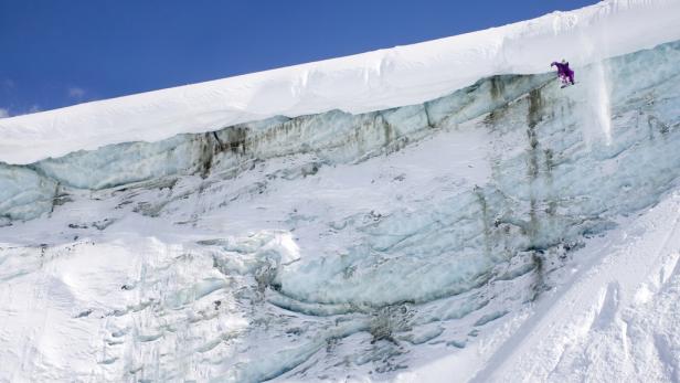 Ein Snowboarder springt von einer schneebedeckten Klippe vor blauem Himmel.