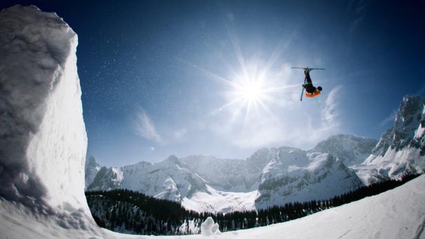 Ein Skifahrer macht einen Rückwärtssalto in der Luft vor einer verschneiten Berglandschaft.