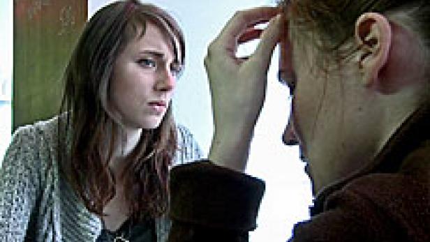 Zwei Frauen sitzen an einem Tisch, eine wirkt besorgt, die andere hält ihre Hand an die Stirn.