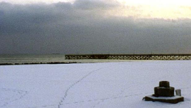Ein schneebedeckter Strand mit einem Pier im Hintergrund unter einem bewölkten Himmel.