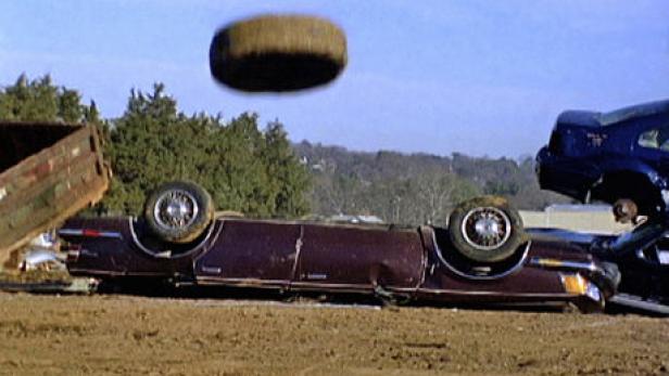 Ein umgedrehtes, beschädigtes Auto auf einem Schrottplatz mit einem fliegenden Reifen.